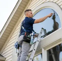 Professional window cleaner on a ladder using a squeegee to clean an exterior arched window, Lowell, MA.